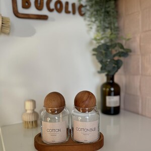 May include: Two clear glass jars with wooden lids labeled "COTTON" and "COTTON BUD" sit on a wooden tray. The jars are on a white surface, with a wooden sign that says "BUCOLIC" in the background. A wooden brush is visible to the left.