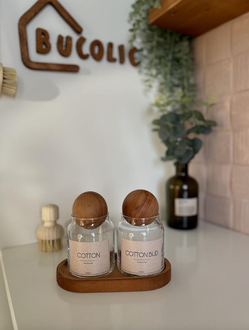 May include: Two clear glass jars with wooden lids labeled "COTTON" and "COTTON BUD" sit on a wooden tray. The jars are on a white surface, with a wooden sign that says "BUCOLIC" in the background. A wooden brush is visible to the left.