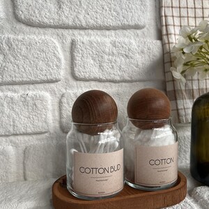 May include: Two glass jars with wooden lids sit on a wooden tray. One jar is labeled "COTTON BUD" and the other "COTTON". Both jars contain cotton swabs. The background features a white brick wall and a checkered towel.