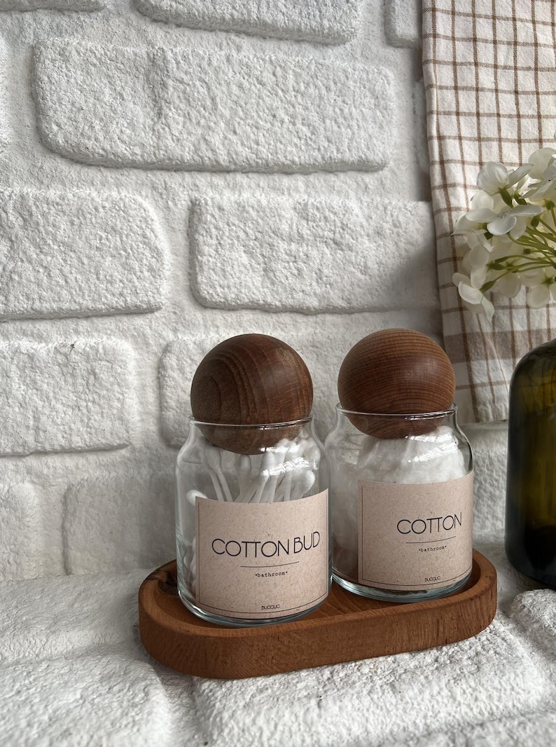 May include: Two glass jars with wooden lids sit on a wooden tray. One jar is labeled "COTTON BUD" and the other "COTTON". Both jars contain cotton swabs. The background features a white brick wall and a checkered towel.