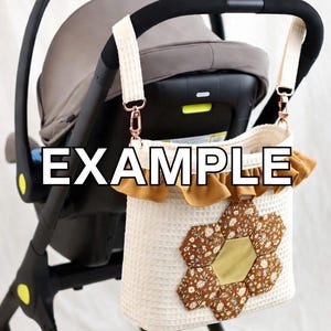以下が含まれることがあります： 花柄のデザインが施された、クリーム色のベビーカーオーガナイザーバッグ。バッグは、クリーム色のストラップとローズゴールドの留め金でベビーカーに取り付けられています。バッグはワッフルテクスチャで、茶色と金の花のアップリケが付いています。
