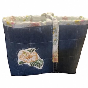 May include: A denim tote bag with a floral embroidered patch. The bag features a quilted design with a floral patterned lining and a matching handle. The embroidered patch showcases a peach-colored hibiscus flower and green leaves.
