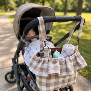 May include: A beige and white gingham fabric stroller organizer bag with a ruffled top edge, featuring two fabric straps with black metal clips for attachment to a stroller handle, containing a water bottle and a rolled patterned cloth.