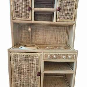 May include: A light-colored, woven rattan play kitchen with a cabinet, stove top, and oven. The cabinet doors and back panel feature a woven design. The knobs and door handles are a dark reddish-brown color. The kitchen has a shelf and a small drawer.