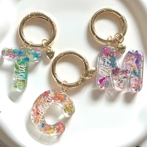 May include: Three gold keyrings with clear resin letter charms. Each charm is filled with pressed flowers and has a name written inside. The names are "Tiana", "Catherine", and "Mary".