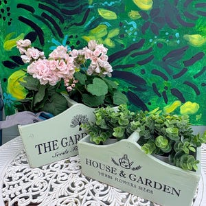 May include: Two light green wooden planter boxes with black text and graphics. One box features the text THE GARDEN and a tree icon, while the other displays HOUSE & GARDEN, HERBS FLOWERS SEEDS, and a watering can icon. Both contain green plants.