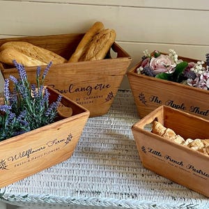 May include: Four wooden baskets with different labels. The baskets are filled with various items, including lavender, bread, and flowers. The labels read "Wildflowers Farm", "Boulangerie", "The Apple Shed", and "Little French Pastries".