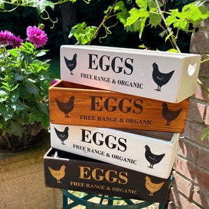 May include: A stack of four rectangular wooden crates for egg storage. Each crate features the text EGGS and FREE RANGE & ORGANIC with black or gold chicken silhouettes. The crates are finished in white, natural wood, white, and dark brown paint.