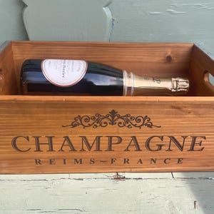 May include: A wooden crate with metal accents, featuring the words "Champagne Reims-France". A bottle of champagne is inside the crate.