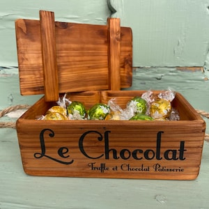 May include: A wooden box with a hinged lid, filled with wrapped chocolates. The box has rope handles and the words "Le Chocolat" and "Truffle et Chocolat Patisserie" are printed on the front in black.