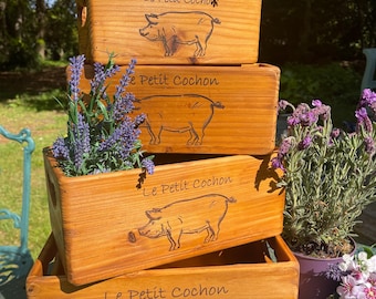 May include: Four wooden planter boxes stacked on top of each other. Each box has a pig design and the text "Le Petit Cochon" engraved on the front. The boxes are filled with lavender plants.