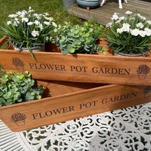 May include: Two wooden rectangular flower pot planters with handles, each holding greenery and white flowers. The words "FLOWER POT GARDEN" are printed on the front of each planter. The planters are sitting on a white table.