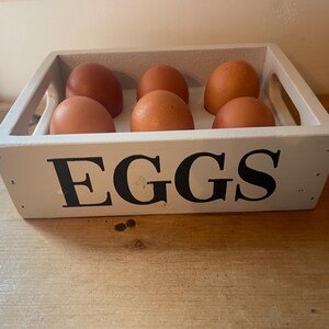 May include: A white wooden egg crate with the word "EGGS" in large black letters. The crate holds six brown eggs. The crate has handles on each side and sits on a wooden surface.