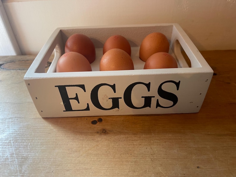 May include: A white wooden egg crate with the word "EGGS" in large black letters. The crate holds six brown eggs. The crate has handles on each side and sits on a wooden surface.