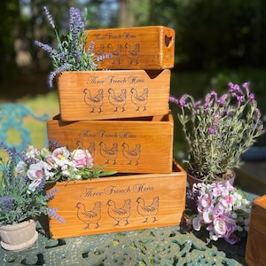 May include: Four wooden boxes stacked on top of each other. Each box is decorated with a design of three French hens and the words "Three French Hens". The boxes are sitting on a green metal table with flowers in front of them.