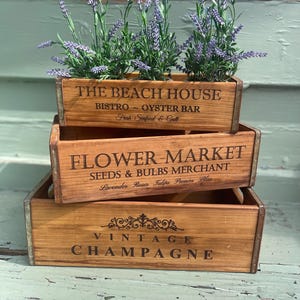 Op de afbeelding: Drie houten kisten op elkaar gestapeld. De bovenste kist heeft de tekst "The Beach House Bistro Oyster Bar Fresh Seafood & Grill". De middelste kist heeft de tekst "Flower Market Seeds & Bulbs Merchant Lavender Roses Tulips Peonies Lilies". De onderste kist heeft de tekst "Vintage Champagne".