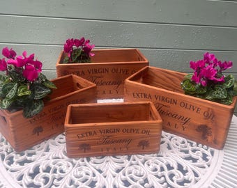 May include: A collection of five wooden rectangular planters, each with a different size. The planters are stained a warm brown and feature the text "EXTRA VIRGIN OLIVE OIL Tuscany ITALY". Each planter holds a vibrant pink flowering plant.