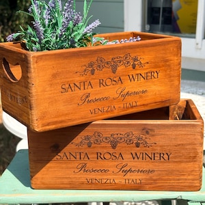 May include: Two rectangular wooden crates with a medium brown finish. Each crate features black printed text reading SANTA ROSA WINERY, Prosecco Superiore, and VENEZIA - ITALY, accompanied by a graphic of grapevines. The top crate contains lavender.