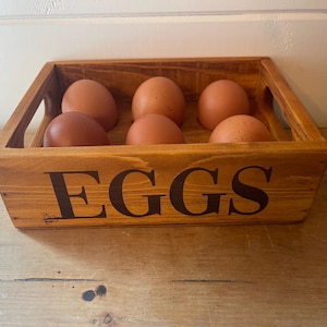 May include: A wooden egg crate with the word "EGGS" in large, dark letters on the front. The crate holds six brown eggs. The crate is made of light-colored wood and has a rustic appearance.