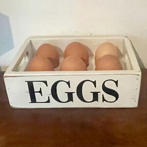 May include: A white wooden egg crate with the word "EGGS" in black lettering. The crate contains several brown eggs. The crate is sitting on a brown surface.