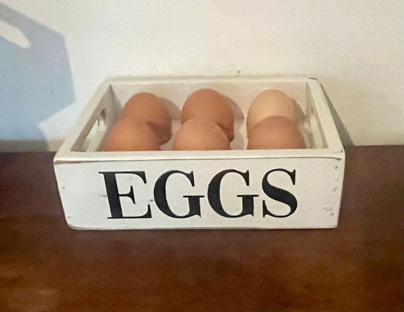 May include: A white wooden egg crate with the word "EGGS" in black lettering. The crate contains several brown eggs. The crate is sitting on a brown surface.