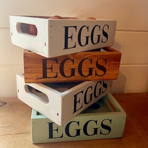 May include: A stack of four wooden egg crates in varying colors: white, brown, and green. Each crate has the word "EGGS" stenciled in black. Some crates contain brown eggs. The crates have cut-out handles.