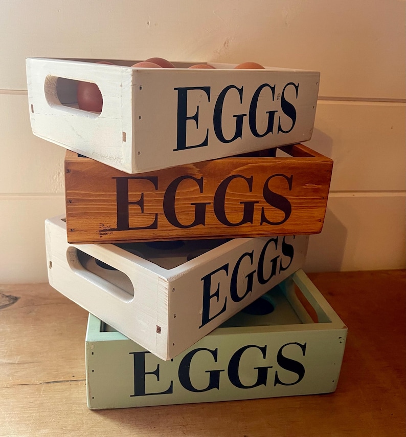 May include: A stack of four wooden egg crates in varying colors: white, brown, and green. Each crate has the word "EGGS" stenciled in black. Some crates contain brown eggs. The crates have cut-out handles.