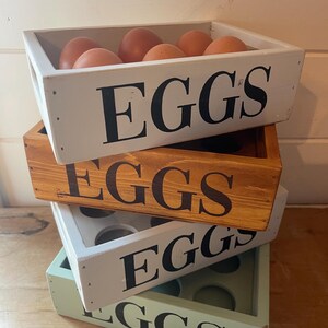 May include: A stack of wooden egg crates in white, brown, and green. The top crate holds brown eggs. Each crate is labeled with the word "EGGS" in large, black letters. The crates are stacked on top of each other.