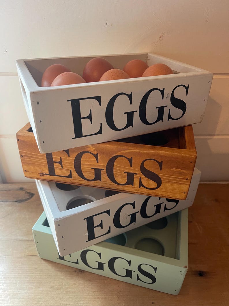 May include: A stack of wooden egg crates in white, brown, and green. The top crate holds brown eggs. Each crate is labeled with the word "EGGS" in large, black letters. The crates are stacked on top of each other.