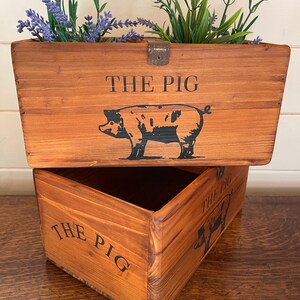 Peut inclure: Un bac à plantes rectangulaire en bois avec le texte "THE PIG" et une illustration de cochon noir. La boîte est remplie de lavande et de verdure. La boîte est en bois avec une finition naturelle.