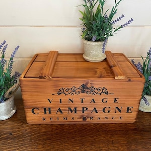 May include: A wooden box with a hinged lid, stained in a warm brown hue. The box is adorned with the words "VINTAGE CHAMPAGNE REIMS FRANCE" in black lettering. Small potted plants with purple flowers flank the box.