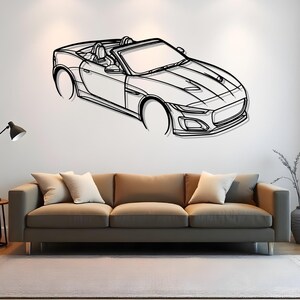 May include: Black and white line art of a convertible sports car mounted on a white wall. The car features a detailed front end, open top, and visible interior. The artwork is displayed above a brown sofa with white cushions.