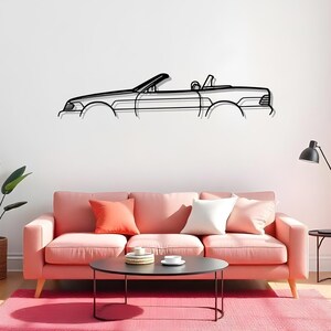 May include: Black metal wall art of a convertible car. The artwork is a line drawing of the car's silhouette, with the top down. The car is mounted on a white wall above a pink sofa with colorful pillows.