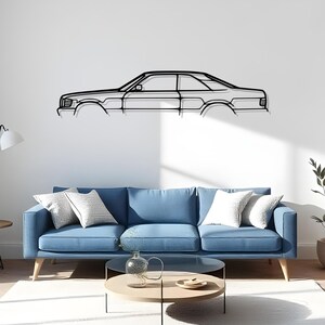 Op de afbeelding: Zwart metalen wandkunst die een zijaanzicht van een klassieke auto afbeeldt. Het kunstwerk is gemonteerd op een witte muur boven een blauwe bank met witte kussens. De kamer heeft een moderne, minimalistische uitstraling.