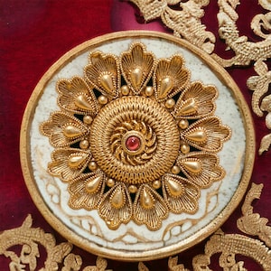 Peut inclure: Un motif floral doré orné d'une pierre précieuse rouge au centre. Le motif est posé sur une base circulaire blanche avec une bordure dorée, sur un fond rouge foncé. Des broderies dorées ornent les bords.