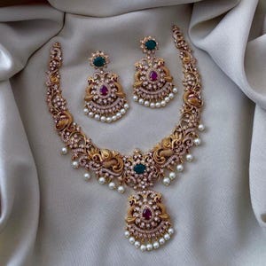 Puede incluir: Un conjunto de collar y pendientes en tono dorado. El collar presenta intrincados adornos, detalles en color esmeralda y rubí, y perlas de imitación colgantes. Los pendientes a juego tienen detalles similares. El conjunto de joyas se exhibe sobre una tela blanca drapeada.
