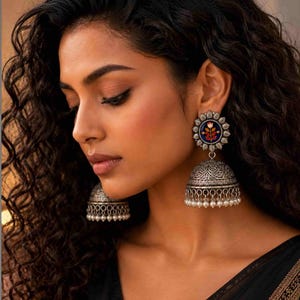 May include: A pair of silver-toned jhumka earrings featuring an oxidized metal finish with intricate engraved patterns. The top circular stud displays a central floral motif in orange, red, and blue, while the bell-shaped base has hanging beads.