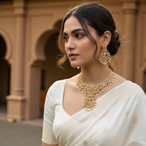 May include: A gold-toned necklace and matching earrings set with intricate detailing and small pearls. The jewelry is worn with a white top. The necklace has a large pendant. The earrings are circular.
