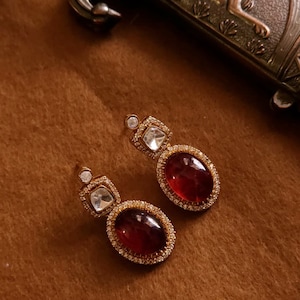 May include: A pair of gold-toned drop earrings. Each earring features a large, oval, deep red gemstone surrounded by small, clear stones. A smaller, square, clear stone is set above the oval stone, with a small, round stone at the top.