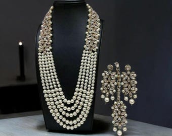 May include: A multi-strand necklace featuring rows of white pearls and clear crystal floral accents, paired with matching drop earrings. The necklace is displayed on a black stand, while the earrings feature a cluster of crystals and pearls.