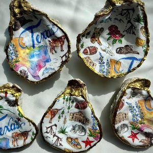 May include: Five oyster shells painted with gold trim and colorful designs depicting Texas landmarks and imagery. The shells are arranged on a white surface. The word "Texas" is written on one of the shells.