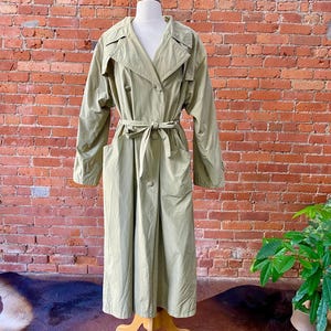 May include: A light green trench coat displayed on a mannequin. The coat features a double-breasted design, a belted waist, and pockets. The coat is long, reaching the ankles, and has a classic trench coat collar. The background is a brick wall.