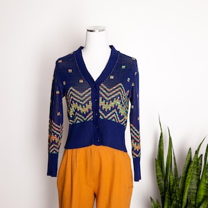 May include: A navy blue cardigan with a V-neck and button closure. The sweater features a geometric pattern in shades of orange, green, and yellow. The cardigan is displayed on a mannequin with orange pants.