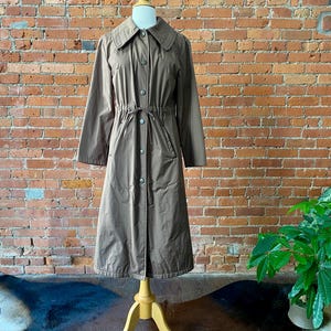 May include: A brown trench coat on a mannequin. The coat has a collared neckline, button-down front, and a drawstring waist. The knee-length coat has long sleeves. The mannequin is on a wooden stand, against a brick wall. The coat's length is approximately 110 cm.