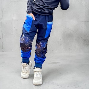Könnte beinhalten: Dunkelblaue Jogginghose mit hellblauen Taschenakzenten und einem Galaxie-Druckmuster an den Knien. Die Hose hat einen dunkelblauen Bund, gerippte Bündchen an den Knöcheln und wird mit einem dunklen Langarmshirt und weißen Turnschuhen getragen.