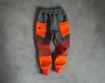 Puede incluir: Pantalones de chándal con bloques de color, base gris oscuro, bolsillos y paneles inferiores en naranja brillante, y una sección central en color óxido. Presentan cintura elástica, puños acanalados en los tobillos y un pequeño parche cuadrado.