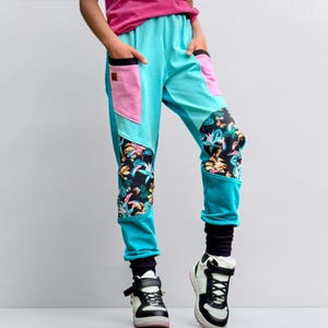 May include: Turquoise joggers featuring pink pocket accents and black panels with a colourful tropical floral print. The trousers have black ribbed cuffs at the ankles and a small brown rectangular patch on the right pocket. The design includes side pockets.