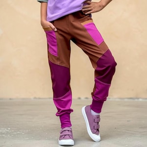 Könnte beinhalten: Farbblock-Jogginghose mit brauner Basis und leuchtend magentafarbenen sowie violetten Einsätzen an Oberschenkeln, Waden und Taschen. Die Hose hat eng anliegende Bündchen an den Knöcheln und wird mit malvenfarbenen Sneakern mit zwei Klettverschlüssen kombiniert.