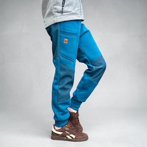 Könnte beinhalten: Hellblaue Jogginghose mit dunkelblauen Einsätzen an Oberschenkeln und Waden, gerippten Beinabschlüssen und einem kleinen quadratischen braunen Aufnäher an der rechten Hüfttasche. Die Hose wird mit braunen Wildleder-Sneakern kombiniert.