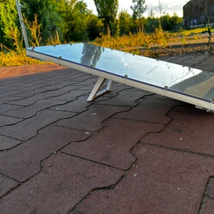 Può includere: Un pannello solare bianco con un top in vetro trasparente è inclinato su una superficie di mattoni rossi. Il pannello è sostenuto da un supporto in metallo bianco.